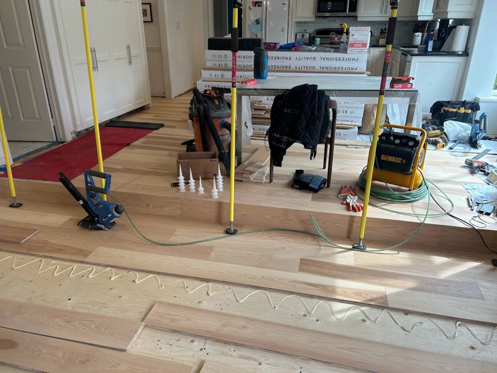 A kitchen with white cabinets with the floor partially torn up and new pieces of wood flooring are being laid. There is glue on the floor where the new wood is about to be placed.